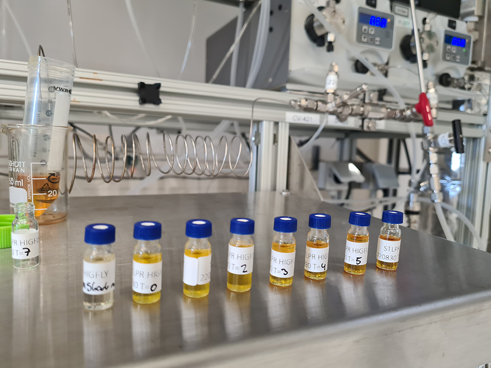 Lab vials with samples on a steel surface, with equipment and tubing in the background.