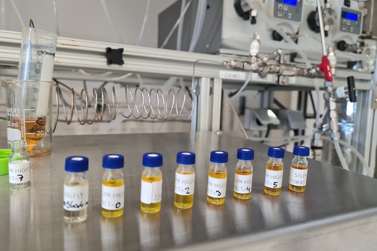 Lab vials with samples on a steel surface, with equipment and tubing in the background.