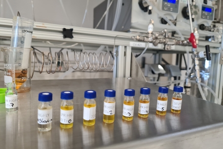 Lab vials with samples on a steel surface, with equipment and tubing in the background.